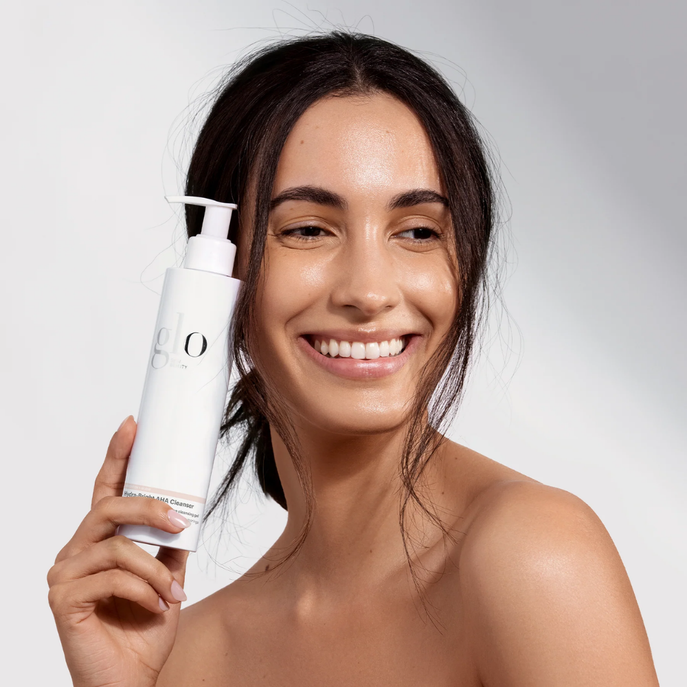 Woman holding a pump bottle of Glo Skin Beauty Hydra-Bright AHA Cleanser with a plain background