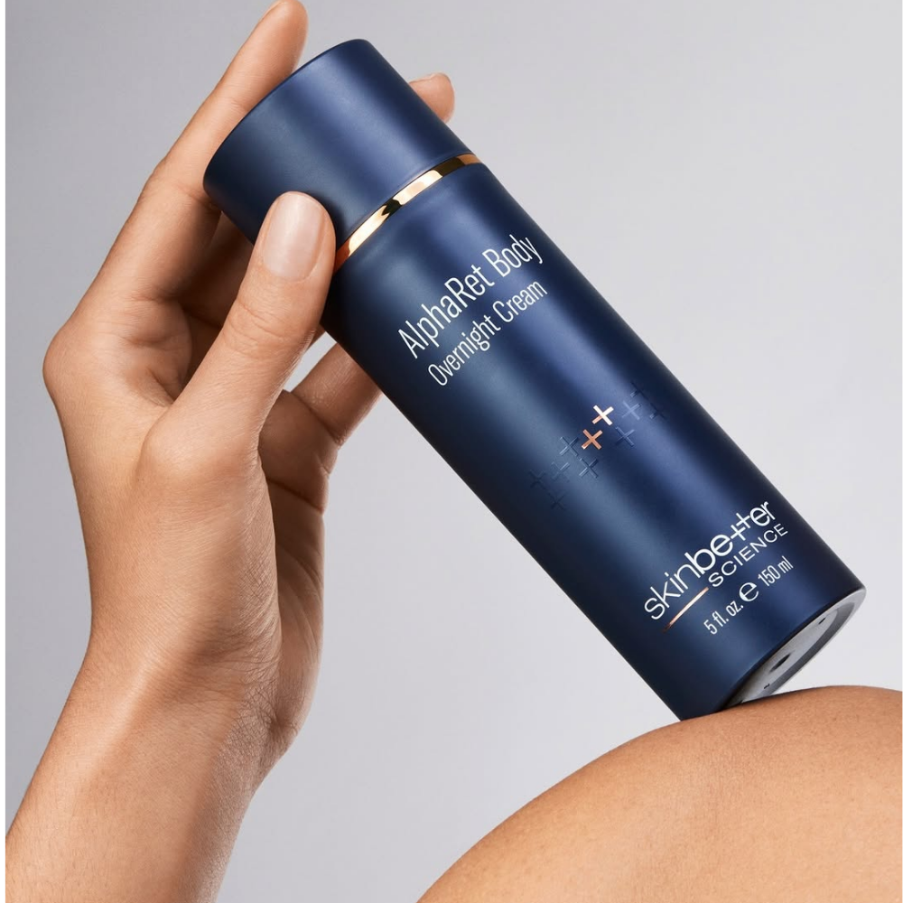 Hand holding a blue skincare cream tube labeled 'Skin Better AlphaRet Body Overnight Cream' on a gray background
