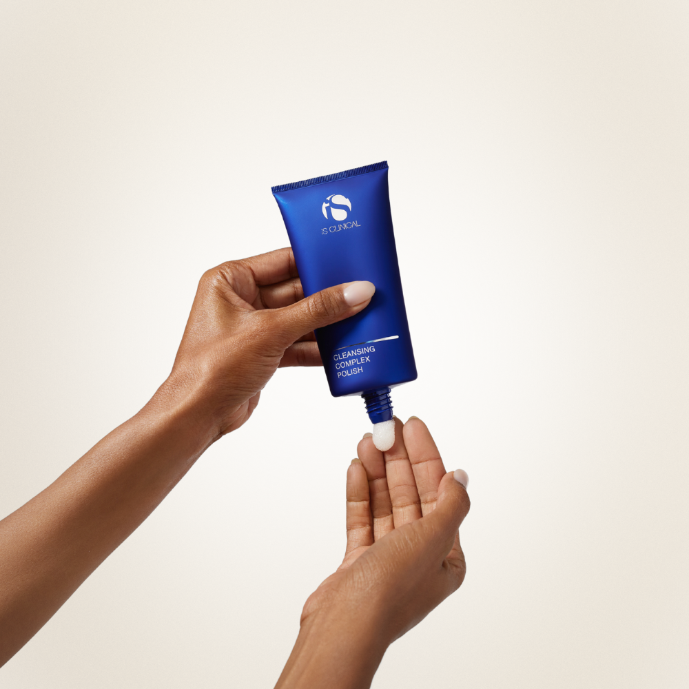 Hand holding a blue skincare tube iS Clinical Cleansing Complex against a beige background