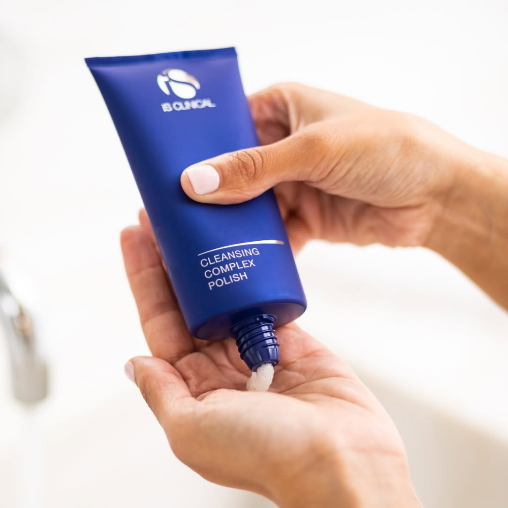 Blue tube of skincare product labeled iS Clinical Cleansing Complex Polish being held with a hand on a white background