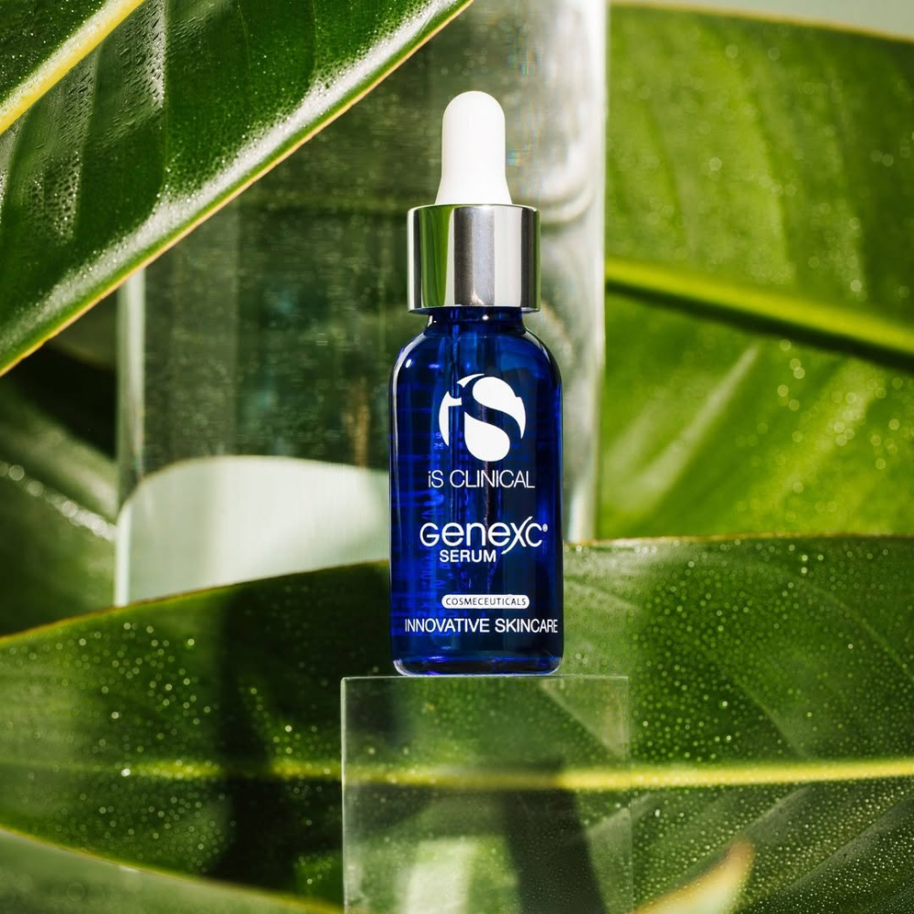 Blue serum bottle labeled iS Clinical GeneXC Serum on a reflective surface with green leaves in the background