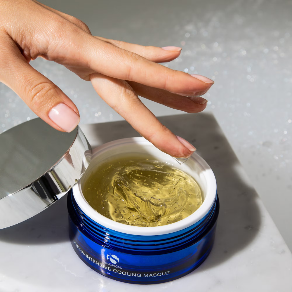 Hand applying a yellow gel from a blue jar labeled iS Clinical Hydra-Intensive Cooling Masque on a gray background