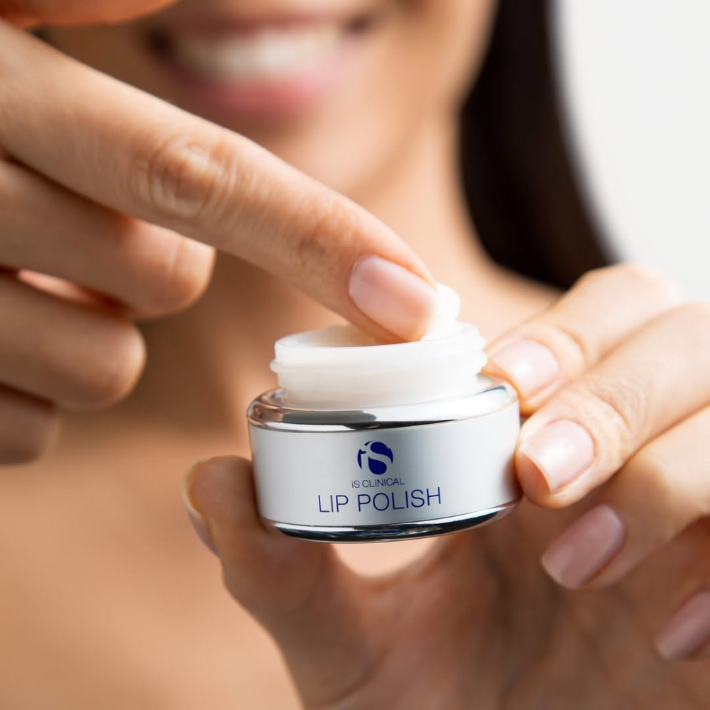 Person applying lip polish from a jar labeled 'Lip Polish' from the iS Clinical Lip Duo with a blurred background