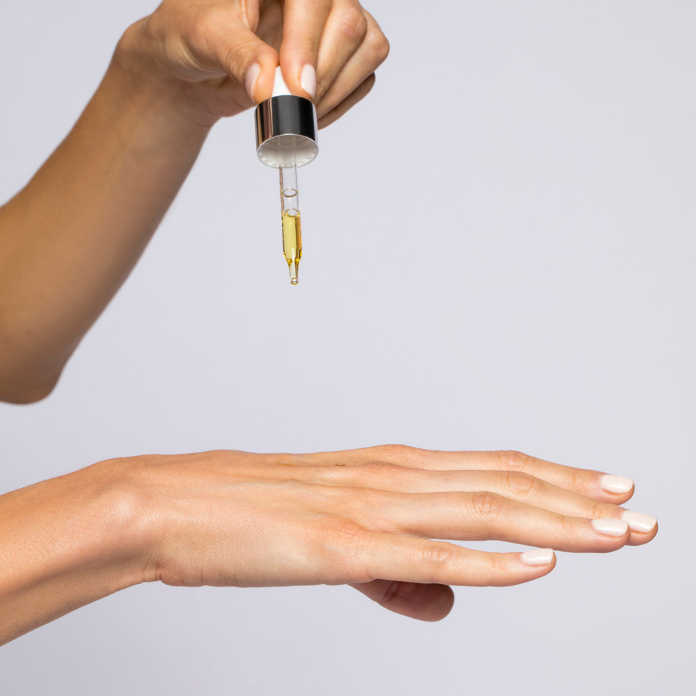 Hand holding a dropper with a small amount of liquid above another hand on a light gray background