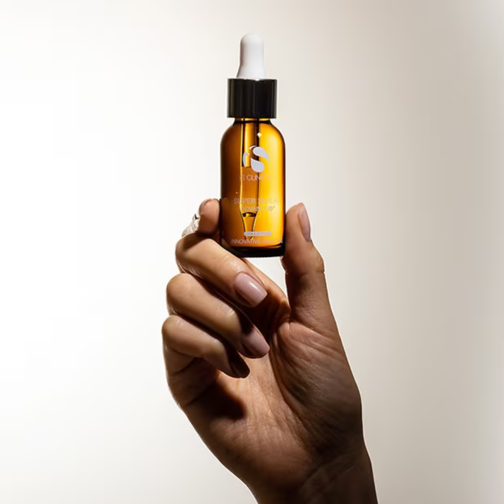 Hand holding an amber glass dropper bottle of iS Clinical Super Serum Advance+ with a white cap against a neutral background