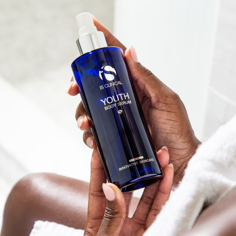 Hand holding a blue bottle of 'Youth Body Serum' by IS Clinical against a neutral background