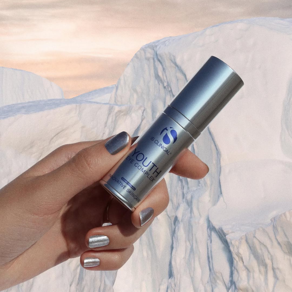 Hand holding a silver skincare bottle labeled iS Clinical Youth Eye Complex against a white rocky background